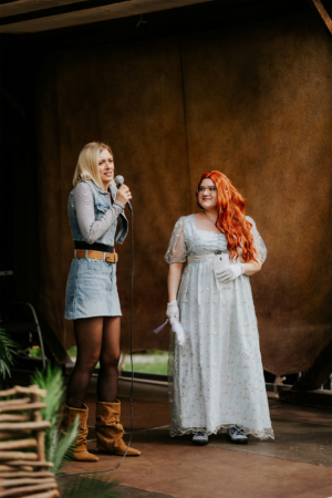 Cosplay Catwalk Zwei Frauen auf der Bühne beim Elbenwald Festival, eine spricht ins Mikrofon, während die andere lächelt. Die erste trägt eine lässige Denim-Jacke und Shorts, die zweite ein elegantes, hellblaues Kleid mit Accessoires. Festivalatmosphäre und kreative Kostüme.