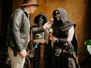 Cosplay Catwalk Ein Moderator interviewt eine verkleidete Teilnehmerin mit Skelett-Make-up und schwarzem Umhang auf dem Elbenwald Festival. Im Hintergrund sind weitere Cosplayer sichtbar, die die kreative Atmosphäre des Events widerspiegeln.