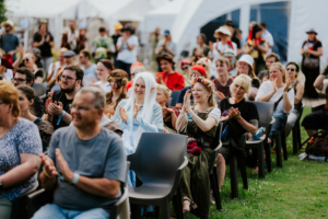 Eine begeisterte Menge applaudiert auf dem Elbenwald Festival, gekleidet in fantasievolle Kostüme. Im Vordergrund sitzen Teilnehmer mit verschiedenen Outfits, während im Hintergrund weitere Festivalbesucher zu sehen sind.