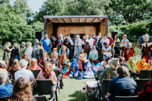 Cosplay Catwalk Bunte Kostüme und kreative Charaktere beim Elbenwald Festival, das eine Mischung aus Musikfestival und Convention bietet. Teilnehmer posieren auf der Bühne, während Zuschauer im Vordergrund gespannt zuschauen. Ein Fest für Nerds und Fantasieliebhaber.
