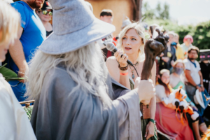 Cosplay Catwalk Ein Szene vom Elbenwald Festival, auf der eine Frau mit Mikrofon einen Zauberer in Kostüm interviewt. Im Hintergrund sind weitere Festivalbesucher in bunten Kostümen zu sehen, die die lebendige Atmosphäre des Events widerspiegeln.