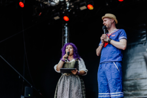 SaKiDe & Olaf Unzel Zwei Moderatoren stehen auf der Bühne des Elbenwald Festivals, umgeben von bunten Lichtern. Die Frau trägt ein historisches Kleid und hält ein Mikrofon, während der Mann in einem maritimen Outfit spricht. Die Atmosphäre ist lebhaft und einladend.