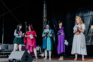 Cosplay Contest Fünf Cosplayerinnen auf der Bühne des Elbenwald Festivals, gekleidet in fantasievolle Kostüme. Sie präsentieren sich vor dem Publikum, während ein Mikrofon und eine Kamera sichtbar sind. Die Atmosphäre ist lebhaft und kreativ, geprägt von der Nerdkultur.