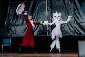 Cosplay Contest Zwei Performer auf der Bühne des Elbenwald Festivals: Eine Frau in einem roten Kleid mit einem Regenschirm tanzt mit einer verkleideten Figur in einem rosa Kostüm und Katzenmaske. Die Atmosphäre ist lebhaft und kreativ, ideal für Fans von Musik und Popkultur.