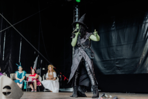 Cosplay Contest Eine Sängerin in einem grünen Kostüm und Hexenhut tritt auf der Bühne auf, während das Publikum in fantasievollen Kostümen zuschaut. Die Atmosphäre des Elbenwald Festivals vereint Musik und Nerd-Kultur in einer einzigartigen Darbietung.