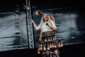 Cosplay Contest Eine performer mit einer kreativen Maske und einem einzigartigen Kostüm steht auf der Bühne des Elbenwald Festivals. Sie hält ein Mikrofon und interagiert mit dem Publikum, während im Hintergrund eine schwarze Bühne sichtbar ist.