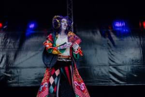 Cosplay Contest Eine bunt kostümierte Person auf der Bühne des Elbenwald Festivals, die mit Karten in der Hand eine Performance präsentiert. Der Hintergrund zeigt eine dunkle Bühne mit bunten Lichtern, die die lebendige Atmosphäre des Festivals unterstreichen.