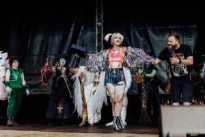 Cosplay Contest Cosplay-Wettbewerb auf dem Elbenwald Festival: Eine junge Frau in buntem Outfit präsentiert sich auf der Bühne, während ein Moderator ihr ein Preis überreicht. Im Hintergrund sind weitere Teilnehmer in fantasievollen Kostümen zu sehen.