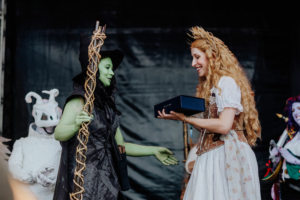Cosplay Contest Eine grün geschminkte Frau in einem Hexenkostüm überreicht einer lächelnden Frau in einem historischen Kleid eine Box auf der Bühne des Elbenwald Festivals, während im Hintergrund weitere verkleidete Teilnehmer zu sehen sind.