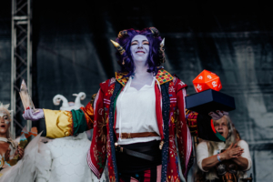 Cosplay Contest Eine cosplayende Person mit lila Haut und Hörnern hält stolz einen Preis und einen roten Würfel auf der Bühne des Elbenwald Festivals, umgeben von jubelnden Zuschauern. Bunte Kostüme und eine festliche Atmosphäre prägen die Szene.
