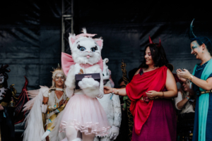 Cosplay Contest Eine Person in einem Katzenkostüm überreicht einen Preis auf der Bühne des Elbenwald Festivals, umgeben von weiteren verkleideten Teilnehmern. Die Atmosphäre ist festlich und bunt, mit kreativen Kostümen und fröhlichen Gesichtern.