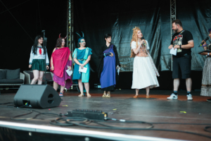 Cosplay Contest Cosplay-Wettbewerb auf dem Elbenwald Festival: Fünf Teilnehmerinnen in kreativen Kostümen stehen auf der Bühne, während ein Moderator Fragen stellt. Die Atmosphäre ist lebhaft und feierlich, umgeben von technischem Equipment und einer wartenden Zuschauermenge.