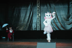 Cosplay Contest Ein Performer in einem Katzenkostüm mit rosa Rock und weißen Stiefeln tanzt auf der Bühne des Elbenwald Festivals, während ein Zuschauer mit einem Regenschirm im Hintergrund sitzt. Die Atmosphäre verbindet Musik und Cosplay in einem bunten Rahmen.