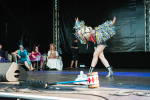 Cosplay Contest Eine Performer*in mit buntem Kostüm führt auf der Bühne des Elbenwald Festivals auf, während eine Gruppe von Zuschauern im Hintergrund sitzt. Die Szene zeigt die kreative Atmosphäre des Festivals, das Musik und Nerdkultur vereint.