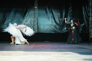 Cosplay Contest Zwei Darsteller auf der Bühne beim Elbenwald Festival, einer verkleidet als geflügelter Engel mit Bogen, der andere als dunkler Charakter mit Trident und dramatischer Kostümierung, präsentieren eine beeindruckende Performance im Rahmen des Festivals.