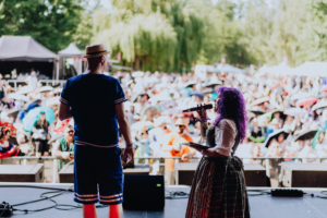 Cosplay Contest Zwei Moderatoren stehen auf der Bühne des Elbenwald Festivals vor einem großen Publikum. Die Frau mit lila Haaren hält ein Mikrofon, während der Mann im blauen Outfit zur Menge spricht. Im Hintergrund sind zahlreiche Zuschauer mit Regenschirmen zu sehen.