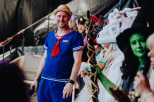 Olaf Unzel Ein lächelnder Mann in einem Matrosenanzug steht auf der Bühne des Elbenwald Festivals, umgeben von verkleideten Teilnehmern in fantasievollen Kostümen. Die Atmosphäre ist lebhaft und feierlich, mit einer Mischung aus Musik und Cosplay.