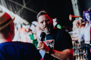 eosAndy Ein Moderator spricht mit einem Mikrofon auf der Bühne des Elbenwald Festivals, umgeben von kostümierten Fans und einer lebhaften Festivalatmosphäre. Die Szene fängt die energiegeladene Mischung aus Musik und Nerdkultur ein.