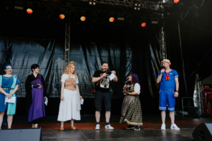 Cosplay Contest Bunt gekleidete Moderatoren und Teilnehmer stehen auf der Bühne des Elbenwald Festivals, während sie ein unterhaltsames Programm präsentieren. Die Atmosphäre ist lebhaft und kreativ, was die Vielfalt der nerdigen Kultur widerspiegelt.