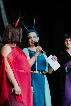 Cosplay Contest Drei Frauen auf der Bühne beim Elbenwald Festival, gekleidet in farbenfrohe Kostüme. Eine Frau mit blauen Haaren und auffälligem Make-up hält ein Mikrofon, während die anderen beiden in roten und dunklen Outfits stehen. Stimmung voller Energie und Kreativität.