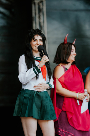 Cosplay Contest Zwei Frauen auf der Bühne beim Elbenwald Festival, eine trägt ein Schulmädchen-Kostüm mit Mikrofon, die andere in einem roten Outfit. Beide lachen und interagieren mit dem Publikum, umgeben von einer festlichen Atmosphäre.