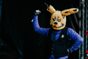Cosplay Contest Ein Teilnehmer des Elbenwald Festivals trägt eine auffällige Maske mit Hasenohren und einem gruseligen Gesicht. Er zeigt seine Begeisterung mit einem kraftvollen Armheben, während er in einem eleganten Outfit mit Weste und Hemd posiert.