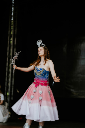 Cosplay Contest Eine Performer*in in einem kreativen Kostüm mit buntem Oberteil und einem glitzernden Rock, umgeben von einer festlichen Atmosphäre auf der Bühne des Elbenwald Festivals, das Musik und Nerd-Kultur vereint.