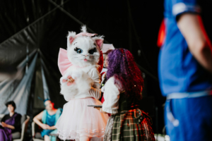 Cosplay Contest Bunt kostümierte Teilnehmer auf der Bühne des Elbenwald Festivals, darunter eine Person in einem großen Katzenkostüm mit Schleife, während eine andere mit lila Haaren ein Mikrofon hält. Im Hintergrund sind weitere Festivalbesucher zu sehen.