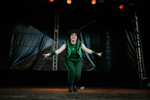 Cosplay Contest Ein fröhlicher Cosplayer in grüner Outfit- und Accessoire-Kombination tanzt auf der Bühne des Elbenwald Festivals. Die energiegeladene Darbietung wird von bunten Lichtern und einer begeisterten Menge unterstützt.