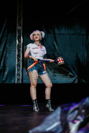 Cosplay Contest Künstlerin auf der Bühne des Elbenwald Festivals, trägt ein T-Shirt mit dem Namen "Harley", gestreifte Shorts und auffällige Stiefel. Sie hält einen bunten Schlagstock und posiert fröhlich vor einem schwarzen Hintergrund.