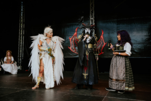 Cosplay Contest Bühne beim Elbenwald Festival mit drei Cosplayern in beeindruckenden Kostümen: ein Engel mit weißen Flügeln, eine dunkle Figur mit roten Drachenflügeln und eine Moderatorin. Die Atmosphäre ist lebhaft und kreativ, perfekt für Fans von Fantasy und Popkultur.