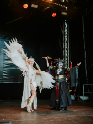 Cosplay Contest Zwei Cosplayer in aufwendigen Kostümen stehen auf der Bühne des Elbenwald Festivals. Die erste trägt ein enges, helles Outfit mit Engelsflügeln und Bogen, während die zweite in einem dunklen, teuflischen Kostüm mit roten Akzenten posiert.