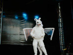 Cosplay Contest Ein fantasievoller Kostümträger in einem weißen Drachenkostüm mit Flügeln steht auf der Bühne des Elbenwald Festivals. Die Hintergrundbeleuchtung sorgt für eine lebendige Atmosphäre, die die kreative und nerdige Stimmung des Festivals unterstreicht.