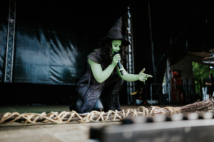 Cosplay Contest Eine grün geschminkte Künstlerin in einem schwarzen Kostüm mit Hexenhut, die auf der Bühne des Elbenwald Festivals ein Mikrofon hält und das Publikum anspricht. Im Vordergrund ist eine dekorative Flechtstruktur sichtbar.