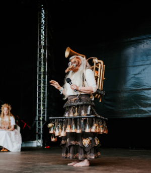 Cosplay Contest Ein Performer in kreativer Kostümierung mit einem hornartigen Gerät auf dem Rücken, steht auf der Bühne des Elbenwald Festivals und hält ein Mikrofon. Im Hintergrund sitzt eine weitere Person in einem fantasievollen Outfit.