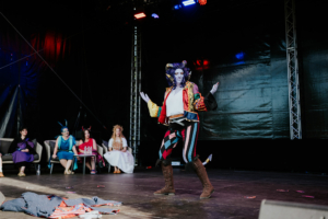 Cosplay Contest Ein Darsteller in auffälligem Kostüm präsentiert eine Performance auf der Bühne des Elbenwald Festivals, während die Jury und Zuschauer im Hintergrund aufmerksam zuschauen. Die bunte Atmosphäre und die kreative Kostümierung spiegeln die Vielfalt des Festivals wider.