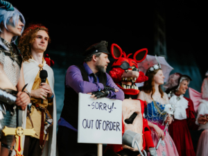 Cosplay Contest Cosplay-Wettbewerb auf dem Elbenwald Festival mit Teilnehmern in fantasievollen Kostümen. Ein Charakter hält ein Schild mit der Aufschrift "Sorry! Out of Order". Die Bühne ist belebt von verschiedenen Figuren aus der Nerd-Kultur, die das Publikum begeistern.