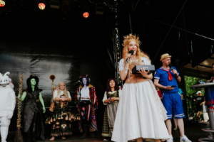 Cosplay Contest Bühne beim Elbenwald Festival mit verschiedenen Charakteren in Kostümen. Eine Moderatorin mit Krone hält ein Mikrofon, während sie von weiteren verkleideten Teilnehmern umgeben ist. Die Atmosphäre ist festlich und kreativ, geprägt von Fantasie und Gemeinschaft.