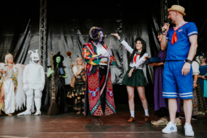 Cosplay Contest Bühne des Elbenwald Festivals mit Teilnehmern in kreativen Kostümen, darunter eine grüne Hexe, ein weißer Drache und verschiedene Charaktere aus Popkultur. Ein Moderator interagiert mit den Darstellern, während das Publikum applaudiert.