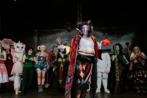 Cosplay Contest Cosplayer auf der Bühne des Elbenwald Festivals, umgeben von anderen Teilnehmern in kreativen Kostümen. Die Hauptperson hält einen Preis und einen roten Würfel, während die Menge applaudiert. Farbenfrohe Kostüme und eine festliche Atmosphäre prägen das Bild.