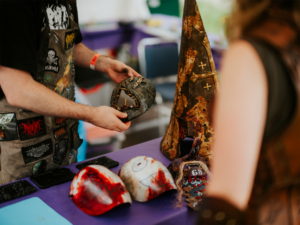 Detailreiche handgefertigte Masken und Kostüme auf dem Elbenwald Festival. Ein Künstler zeigt eine Maske mit einzigartigen Verzierungen, während im Hintergrund weitere kreative Werke auf einem lila Tisch präsentiert werden.