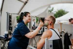 Crafting Village Ein Makeup-Künstler trägt Glitzer auf den Bart eines Mannes auf, während er in einem Stuhl sitzt. Im Hintergrund sind Spiegel und weitere Festivalbesucher zu sehen. Die Szene findet während des Elbenwald Festivals statt, das Musik und Nerdkultur vereint.