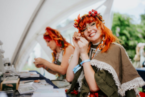 Crafting Village Lächelnde Teilnehmerin mit rotem Haar und Blumenkranz zeigt stolz ein handgefertigtes Kunstwerk auf dem Elbenwald Festival. Im Hintergrund arbeitet eine weitere Person an einem Tisch voller Bastelmaterialien. Festliche Atmosphäre und kreative Gemeinschaft.