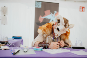 Crafting Village Ein Teilnehmer des Elbenwald Festivals trägt eine Tiermaske und arbeitet an einem kreativen Projekt am Tisch. Um ihn herum sind Bastelmaterialien und technische Geräte zu sehen, die die kreative Atmosphäre des Festivals unterstreichen.