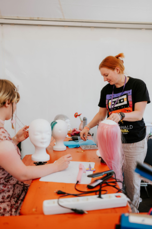 Crafting Village Eine kreative Werkstatt beim Elbenwald Festival, in der Teilnehmer an Perücken arbeiten. Eine Person mit rotem Haar zeigt eine rosa Perücke, während eine andere an einem Kopfmodell arbeitet. Verschiedene Werkzeuge und Materialien sind auf dem Tisch verteilt.