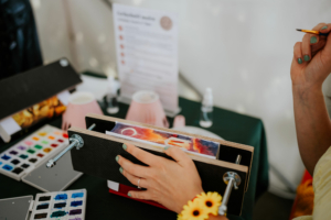 Crafting Village Eine Person malt mit Aquarellfarben an einem Tisch auf dem Elbenwald Festival, umgeben von Malutensilien und einem Informationsschild. Die Hand ist mit einem gelben Armband geschmückt, während die andere Hand den Malblock hält.