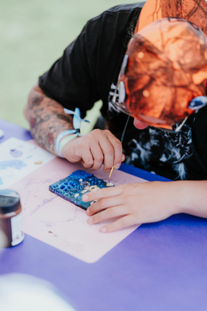 Crafting Village Ein Teilnehmer des Elbenwald Festivals malt konzentriert an einem Kunstprojekt mit blauen und goldenen Farben. Auf dem Tisch sind verschiedene Malutensilien und ein kreatives Design sichtbar, das die künstlerische Atmosphäre des Festivals widerspiegelt.