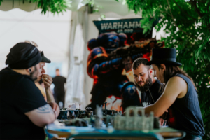 Dice Club Cottbus Vier Männer spielen an einem Tisch mit Miniaturfiguren, umgeben von grünen Pflanzen und einem Warhammer 40k-Plakat im Hintergrund. Die Szene fängt die lebendige Atmosphäre des Elbenwald Festivals ein, wo Gaming und Gemeinschaft im Vordergrund stehen.