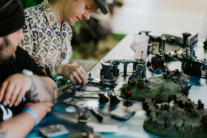 Dice Club Cottbus Zwei Teilnehmer des Elbenwald Festivals konzentrieren sich auf ein Tabletop-Spiel, umgeben von detaillierten Miniaturen und Spielkarten. Der Tisch ist mit einer fantasievollen Landschaft dekoriert, die die kreative Atmosphäre des Festivals widerspiegelt.