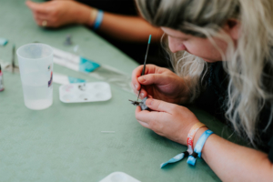 Dice Club Cottbus Eine Teilnehmerin des Elbenwald Festivals malt konzentriert an einer Miniaturfigur, umgeben von Malutensilien und einer Wasserquelle. Die Szene zeigt die kreative Atmosphäre des Festivals, das Musik und Nerdkultur vereint.